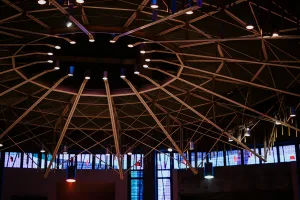 Large circular ceiling framework of radial metal beams and crisscrossing supports studded with downward spotlights and pendant lamps above a dim interior lit by blue-tinted stained-glass windows.