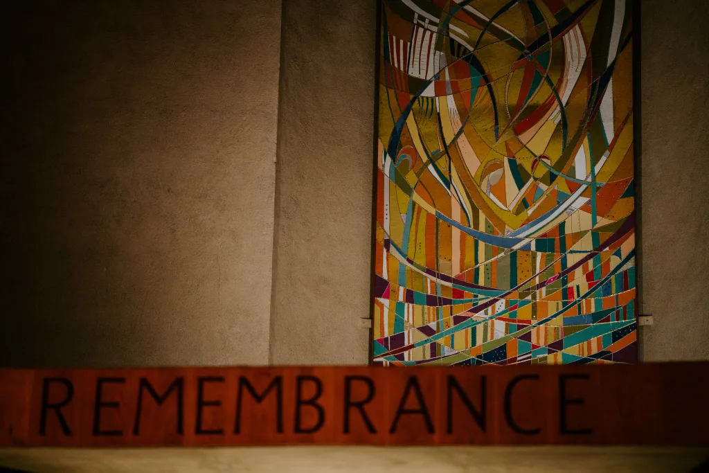 Colourful abstract panel with sweeping curved lines and multicoloured stripes mounted on a plain beige wall, with a wooden plaque in the foreground bearing the word REMEMBRANCE