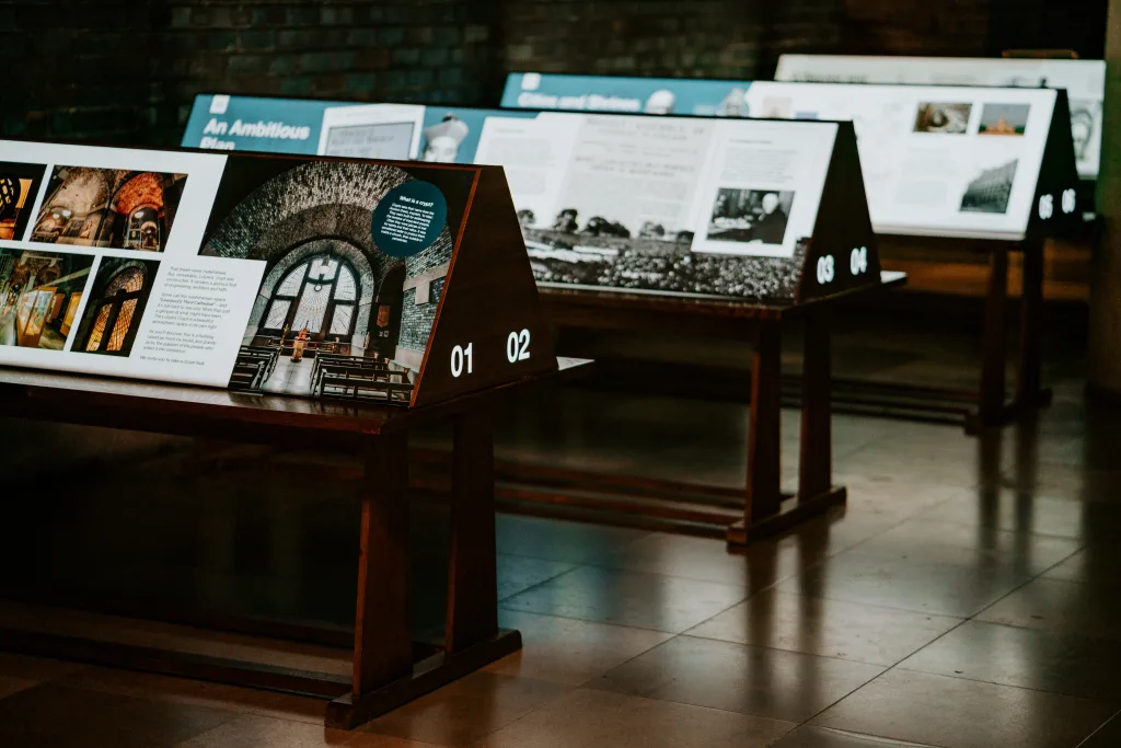 Angled museum display panels on wooden stands showing historical photographs and explanatory text—numbered 01–04—arranged in a dim gallery with a tiled reflective floor and a brick wall background.