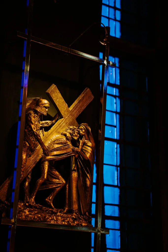 Bronze relief panel depicting Jesus bent under a heavy wooden cross, a mourning woman supporting him and a soldier behind, lit warmly against a tall blue stained-glass window