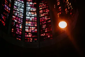 Tall segmented stained-glass windows in reds, purples and oranges with black leaded geometric patterns filling a dark interior, and a bright round hanging light casting a warm glow to the right.