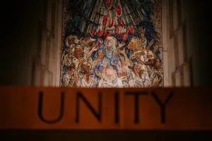 Vibrant mosaic in a church depicting robed figures with raised hands and tongues of flame above their heads, framed by dim stone walls and seen over a blurred wooden panel engraved with the word UNITY.