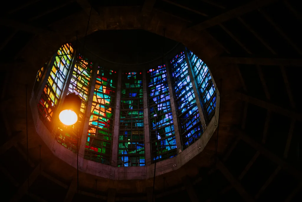 Octagonal stained-glass skylight set into a dark concrete ceiling, with tall vertical panels shifting from warm yellows and greens on the left to deep blues on the right and a glowing hanging pendant lamp near the left edge.