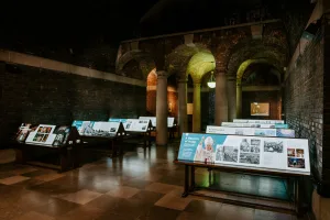 Dimly lit vaulted brick exhibition hall with stone columns and arched ceilings, tiled floor and several illuminated angled information panels on wooden stands, one panel prominently showing a robed religious figure raising a hand.