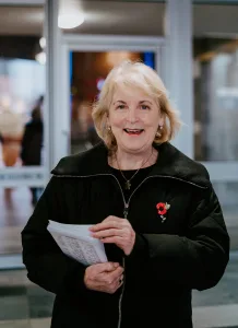 Smiling elderly woman with short blonde hair wearing a black zipped coat and a red poppy brooch, holding a stack of papers in a bright indoor entrance or lobby.