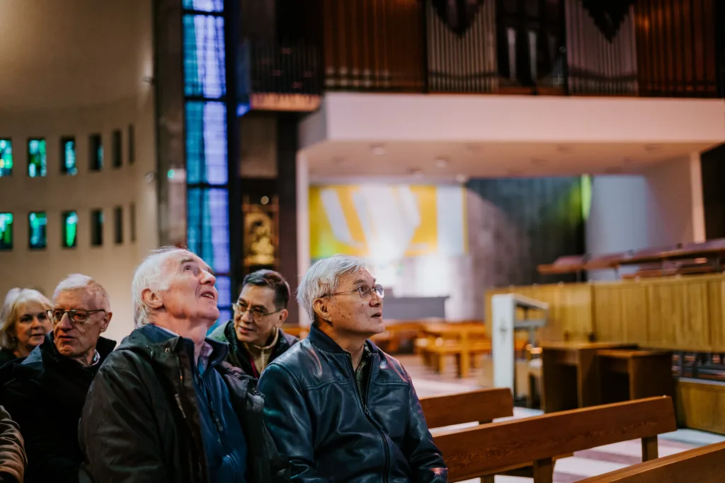 Several older adults seated on wooden pews inside a church, two men in the foreground wearing jackets looking up attentively toward stained-glass windows and organ pipes above.