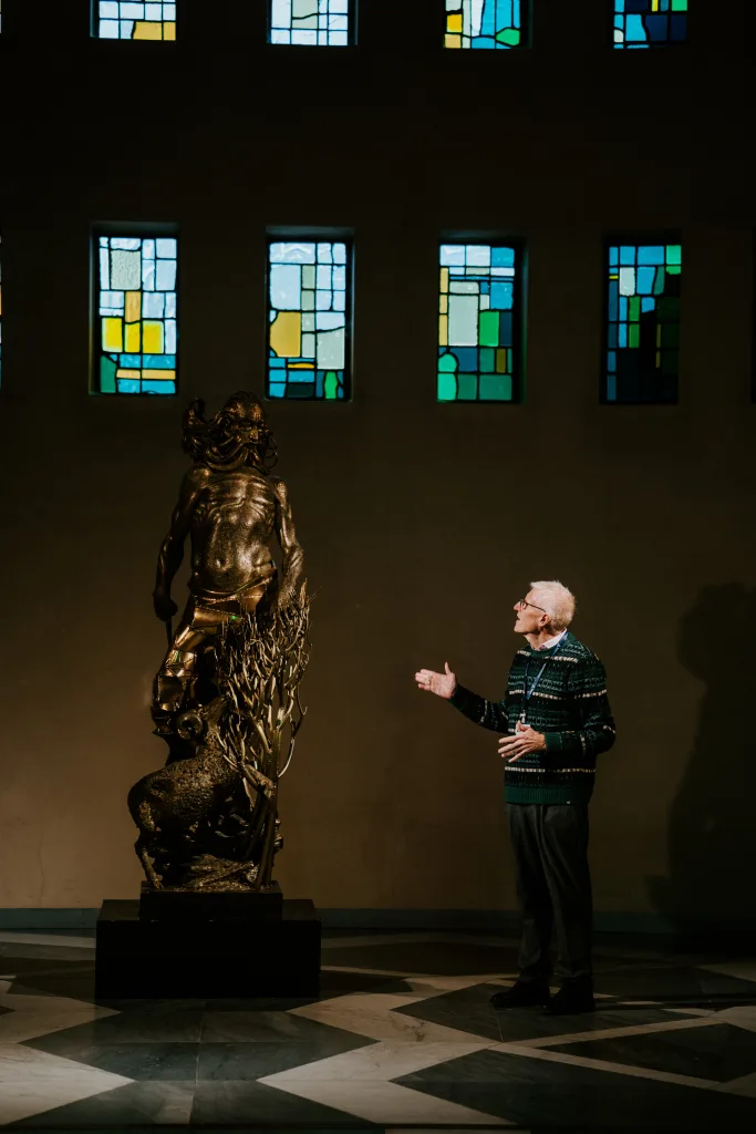 An elderly man in a patterned sweater and glasses gestures toward a tall bronze sculpture of a bearded, muscular figure rising above a ram and twisted branches in a dim gallery lit by a row of small colourful stained-glass windows.