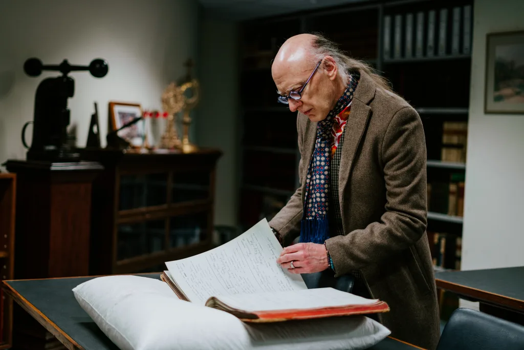 A balding man wearing glasses, a patterned scarf and a brown coat leans over an open historical ledger resting on a white cushion in a dimly lit archival reading room.