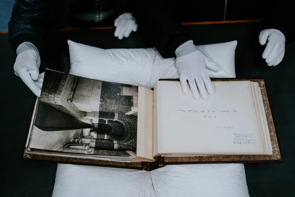 White‑gloved hands carefully turn the pages of an open vintage photographic album resting on a white cushion, revealing a black‑and‑white image of brick arches and a facing page with handwritten notes.