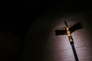 Gold-toned wooden crucifix of Christ with outstretched arms lit by a single spotlight against a textured brick wall, the cross and figure casting a stark shadow into surrounding darkness.