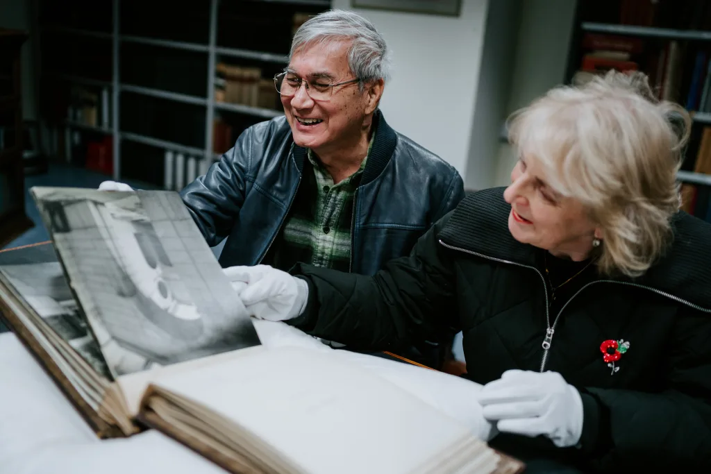 An older man with glasses and an older woman wearing a red poppy pin, both in white archival gloves, smile as they examine a large black-and-white photograph in an open album in a library or archive with bookshelves behind them.
