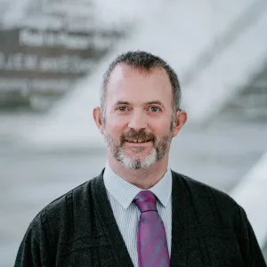 Middle-aged man with short salt-and-pepper hair and a trimmed beard, smiling at the camera while wearing a striped shirt, purple tie and dark cardigan against a soft-focus modern architectural background.