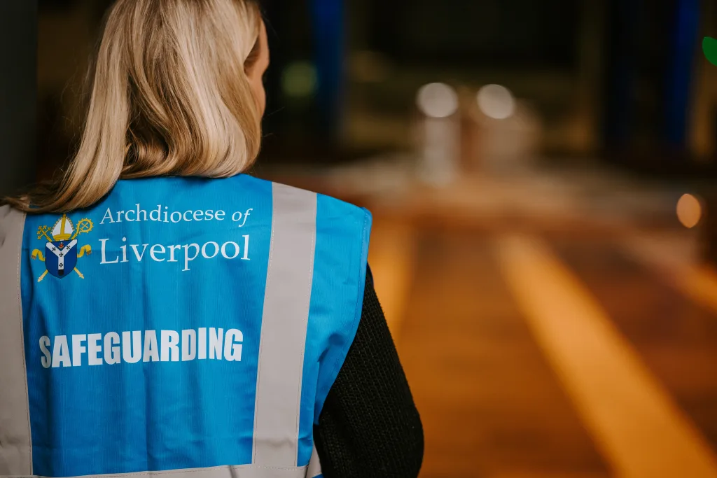 Rear view of a blonde person wearing a blue high-visibility vest printed with the Archdiocese of Liverpool crest and the word SAFEGUARDING, standing in a softly lit, blurred interior.
