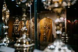 Ornately embroidered ecclesiastical vestments in a glass display case, centered on a gold chasuble with a large embroidered cross, seen through blurred, sparkling silver chalices in the foreground.