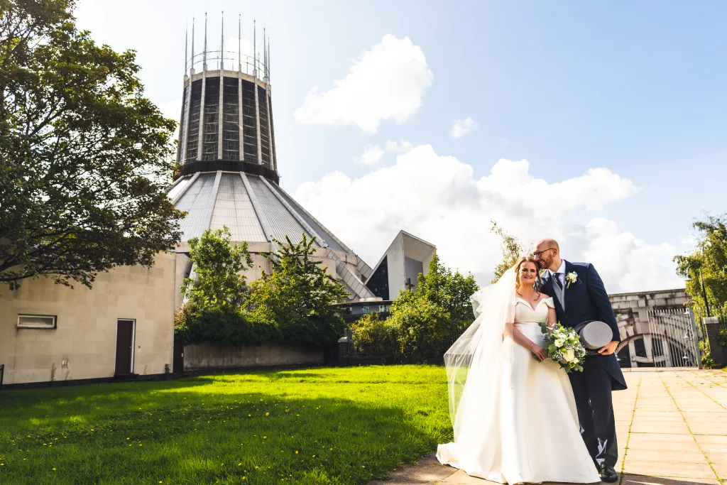Smiling bride in a white gown and veil holding a bouquet stands arm-in-arm with the groom in a dark suit holding a top hat as he kisses her temple, posed on a sunlit path with green lawn and trees in front of a distinctive modern circular church beneath a blue sky with fluffy clouds.