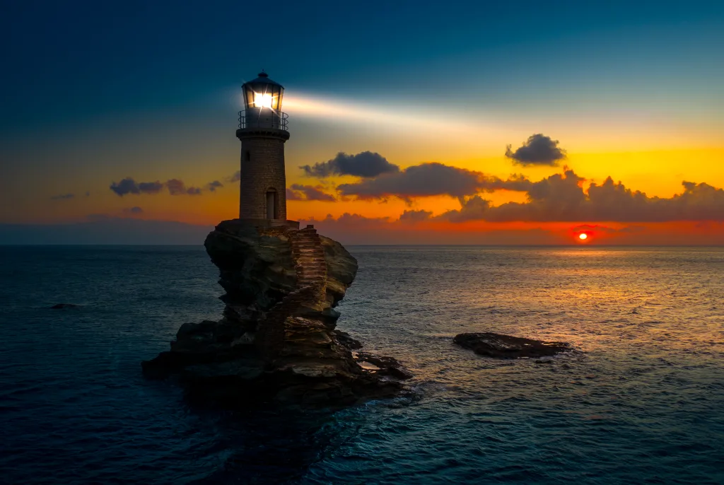 A solitary stone lighthouse crowns a jagged rock outcrop with a winding stone stairway, its bright beacon sweeping across the calm sea as the sun sets behind low clouds on the horizon.