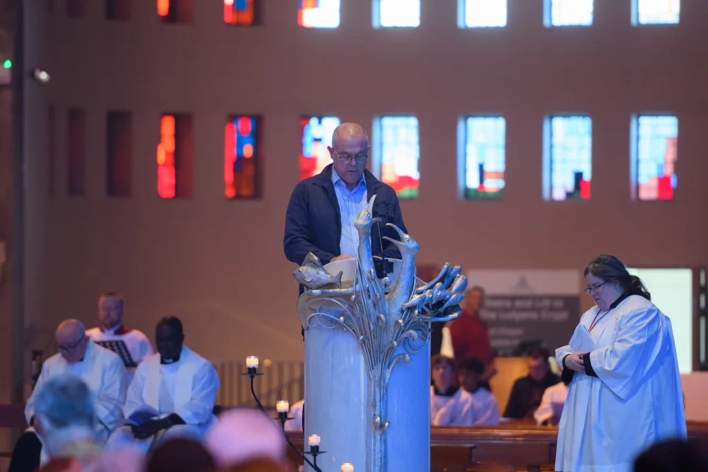 A man reads from an ornate stone lectern decorated with sculpted fish and flowing motifs in a dim church interior lit by colourful stained-glass windows, while clergy and altar servers stand and sit nearby.