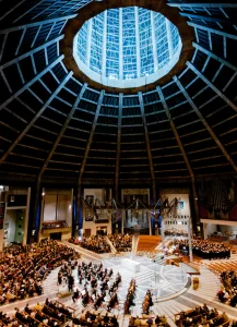 Modern circular cathedral interior with a high ribbed concrete dome and glazed central oculus, an orchestra arranged on a round stage at the centre and a large audience seated in concentric tiers around it.