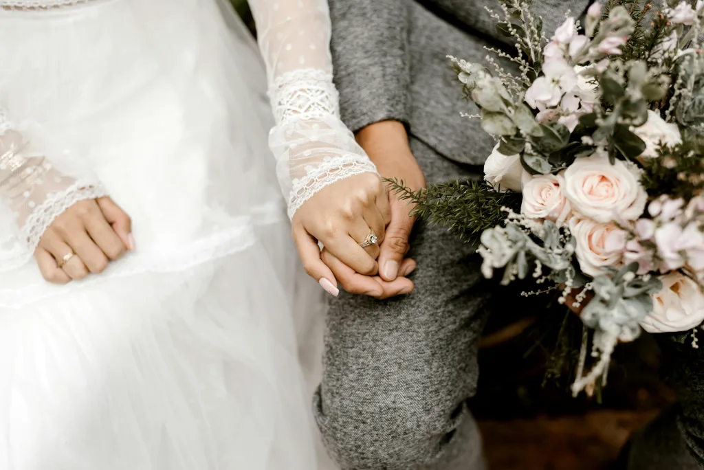 Close-up of a bride and groom holding hands, the bride in a white lace-sleeved dress wearing a diamond ring and the groom in a grey suit beside a bouquet of pale pink roses and greenery.