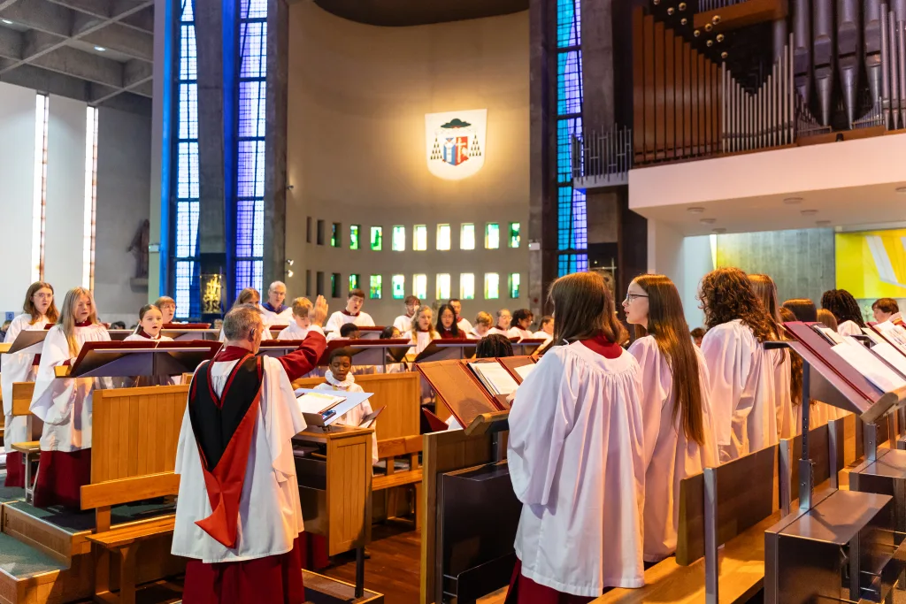 A mixed-age choir in white robes with red collars sings from wooden music stands as a conductor raises his hand in a church with tall blue stained-glass windows and a large pipe organ behind them.