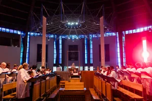 Choir of adults and children in white cassocks stand in opposing wooden stalls, singing as a conductor raises his hands at a central altar beneath a circular metal light rig in a spacious church with tall blue stained-glass windows and a bright red light flare at the right.