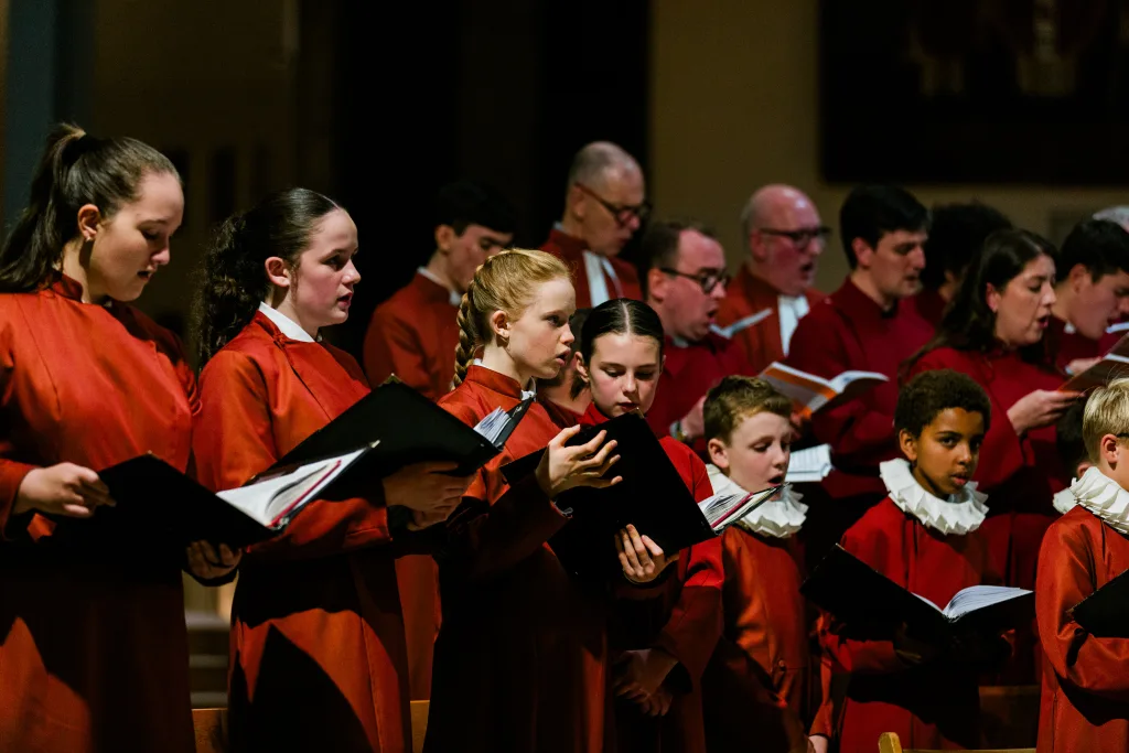 Choir of children and adults in red robes, some boys wearing white ruffled collars, standing in rows in a dim church interior and holding black music folders while singing.