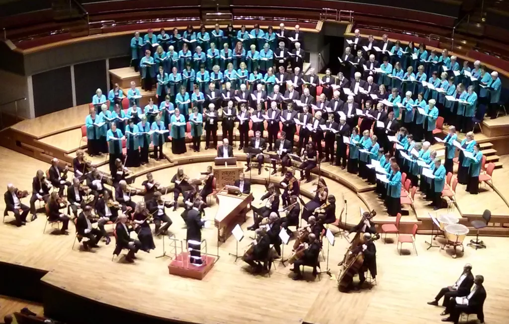 Large choir in teal robes and black formal wear on tiered risers behind a seated orchestra of string and wind players with a conductor on a wooden concert-hall stage.