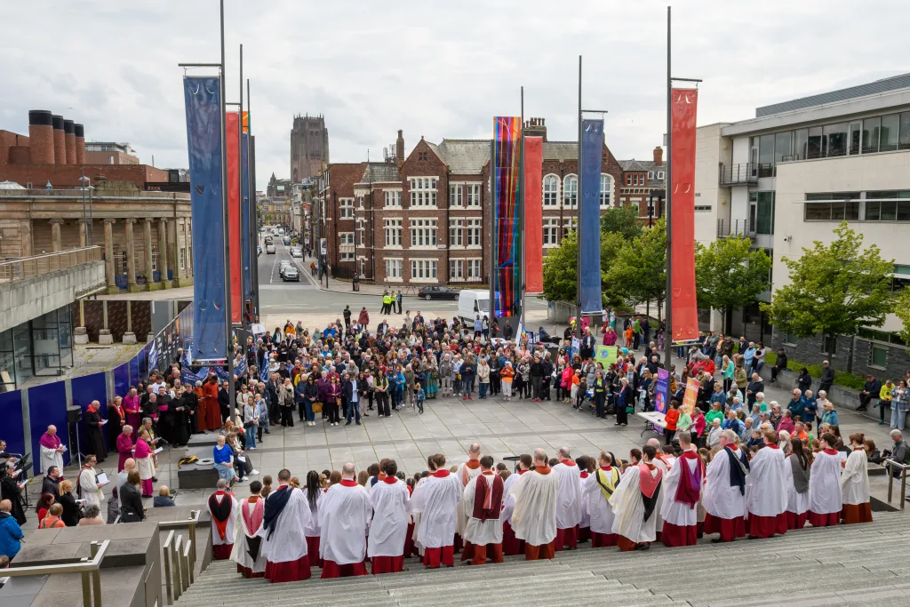 Choir dressed in white surplices over red cassocks stands on wide stone steps singing to a large crowd gathered in a city square framed by tall colourful banners, red‑brick buildings and a distant cathedral tower under a grey sky.