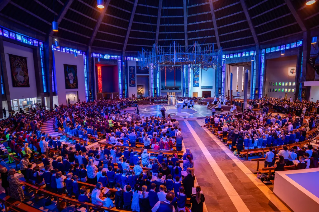 Wide interior view of a large circular modern cathedral filled with a congregation — many children in school uniforms in the foreground — with blue-lit stained-glass windows encircling the space, a central altar beneath a large suspended metal chandelier, and clergy and choir gathered for a service.