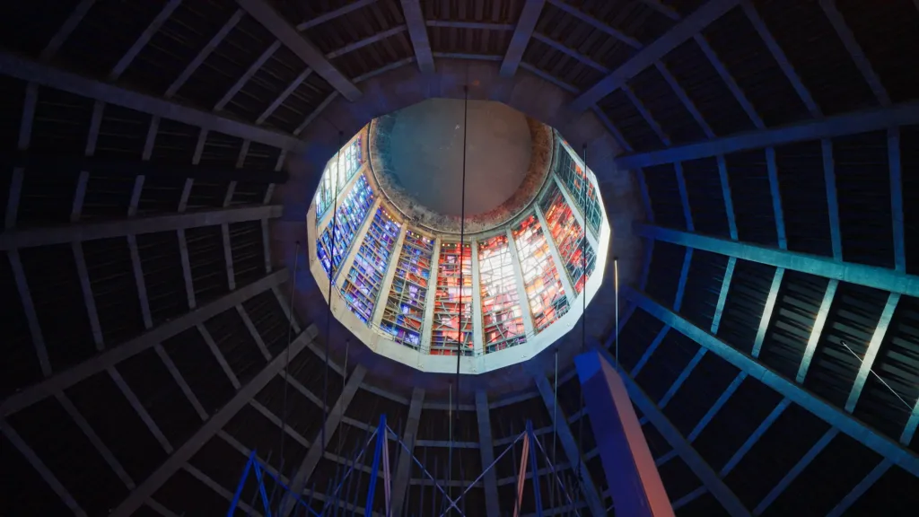 Interior view looking up at a circular concrete dome with an octagonal ring of colourful red and blue stained-glass panels around a central oculus, framed by dark radial ribs.