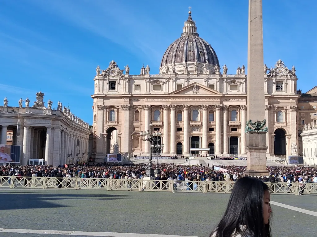 St. Peter's Basilica's grand façade and dome rising behind the Vatican obelisk, with large crowds in St. Peter's Square